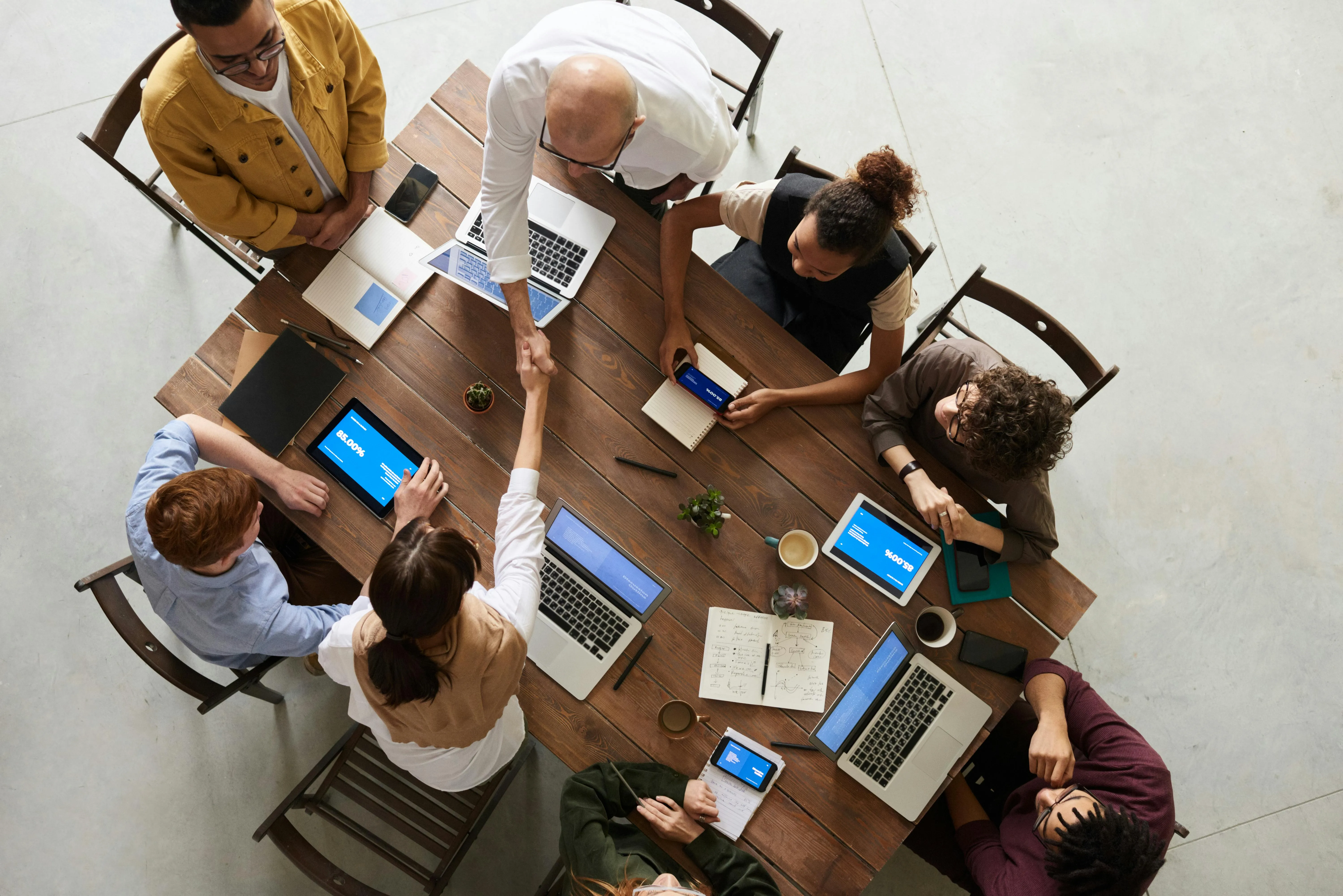 A birds eye view of 8 people sat around a table with laptops, tablets and notepads. 2 people are shaking hands over the table.