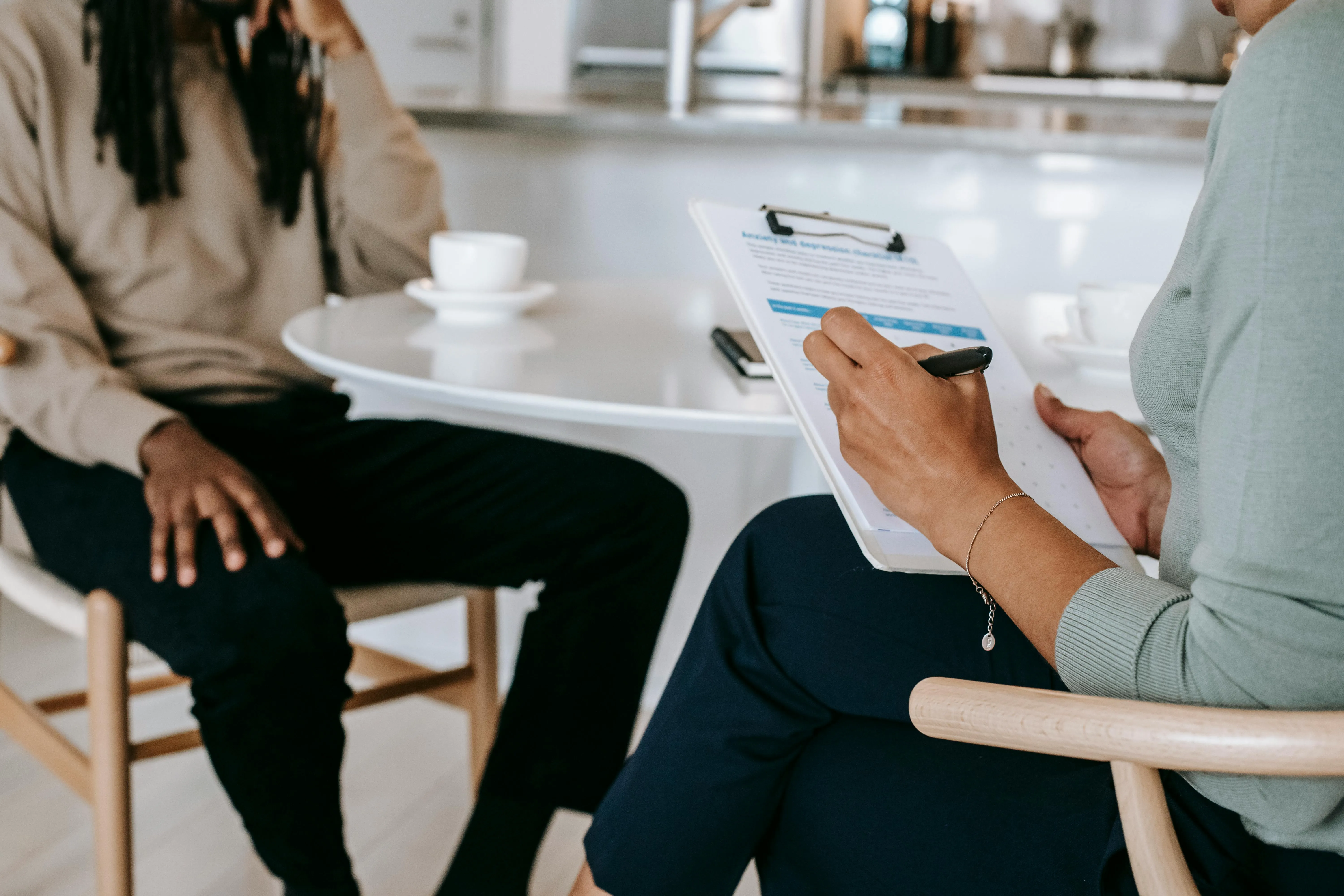 2 people sat at a coffee table with coffee cups, one person has a clipboard and a pen while speaking to the other person.