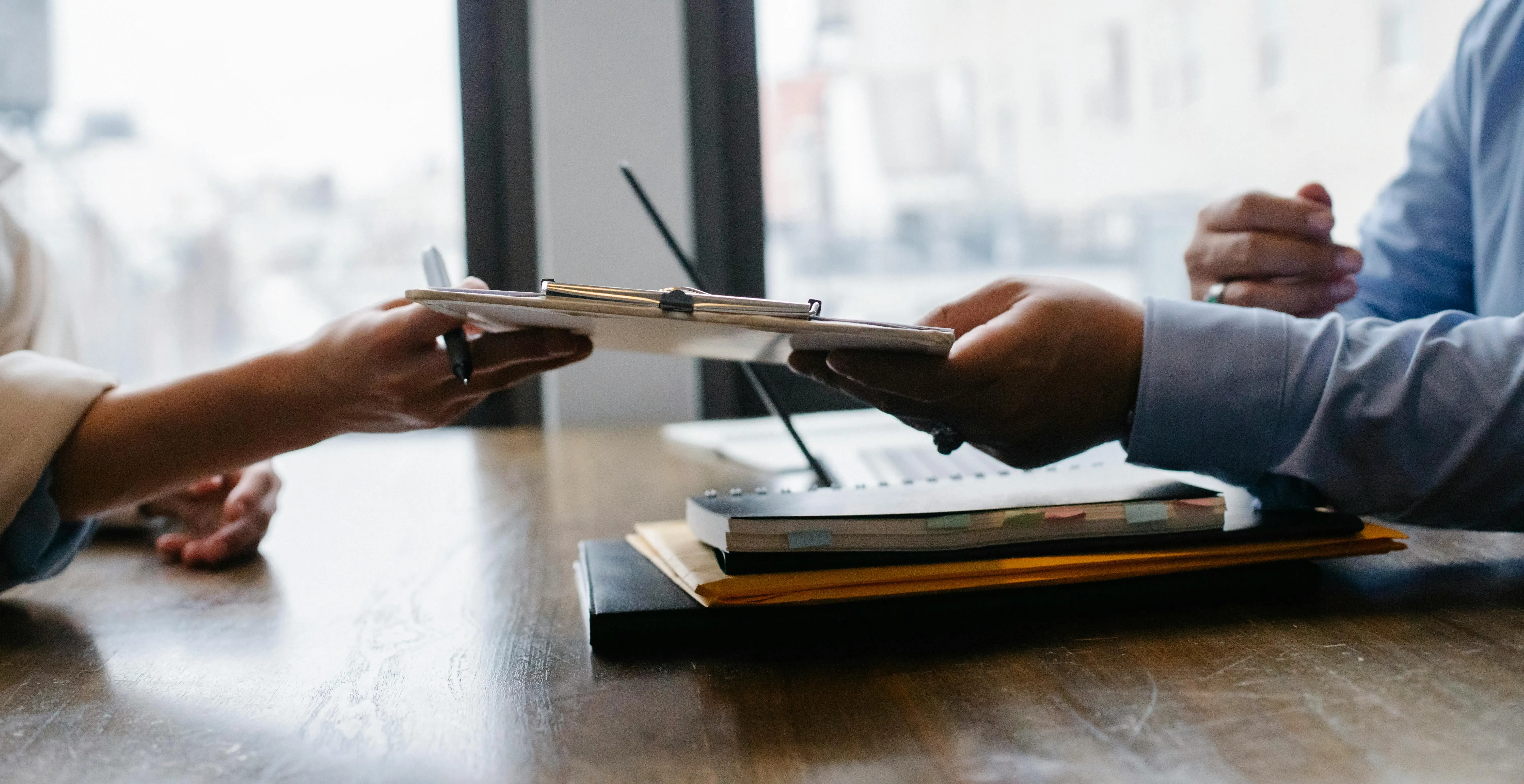 A person handing a clipboard to another person across a table with books on.