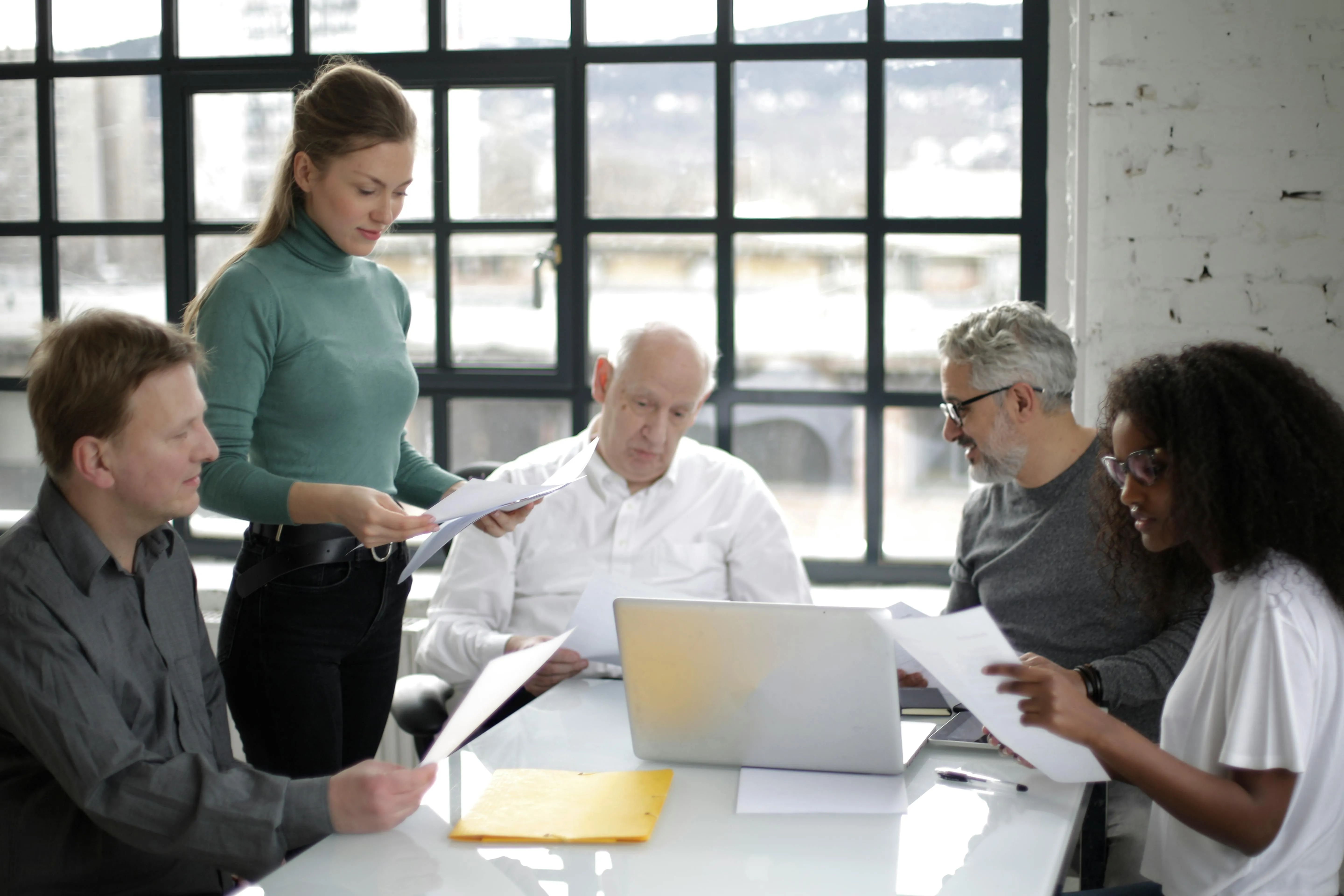 5 people sat around a table with a laptop on it, looking at papers while talking, one person is standing.