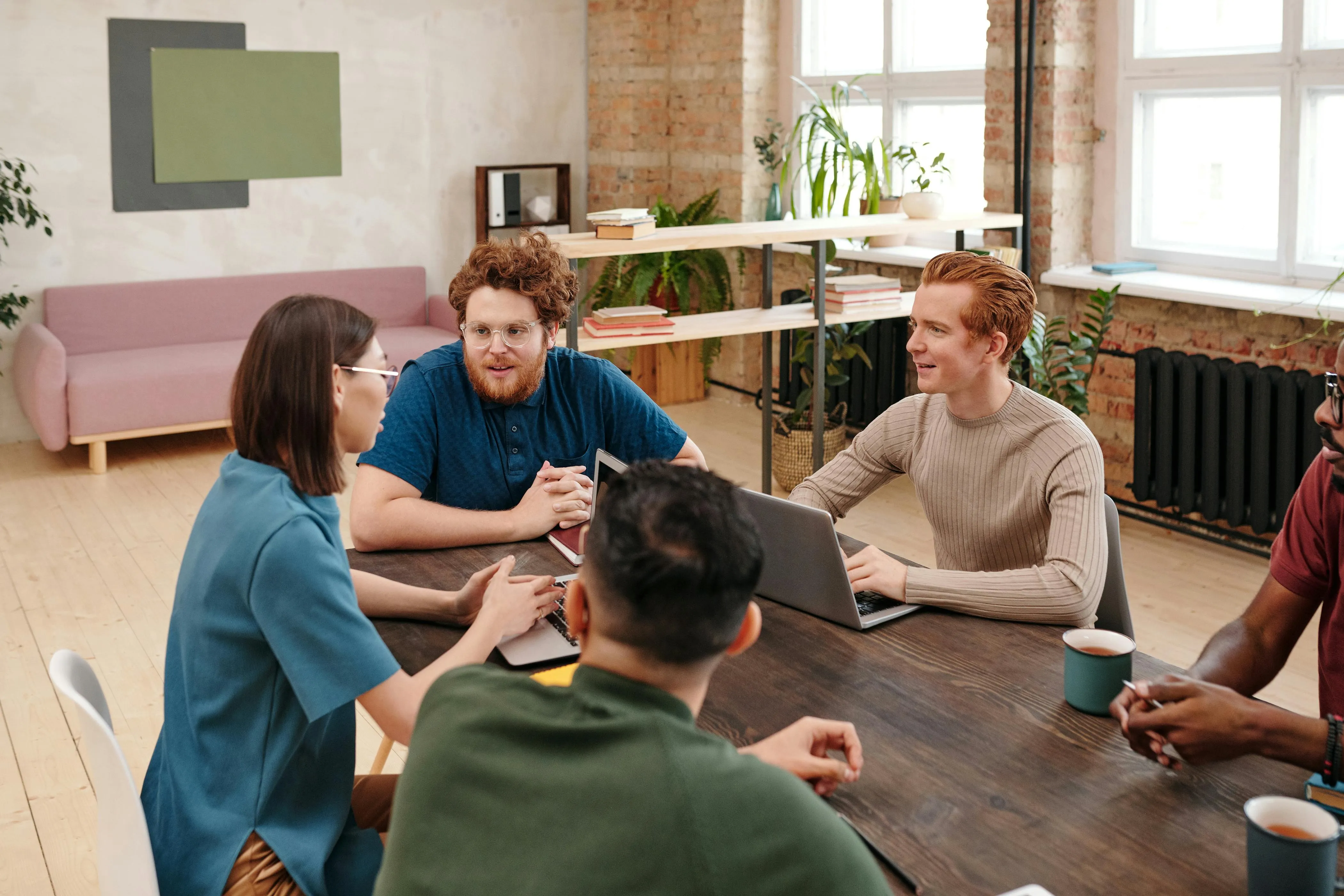 5 people sat around a wooden table all talking with laptops and coffee cups in a large casual workspace.